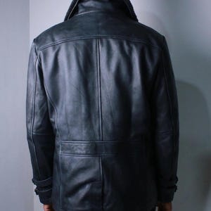 May include: A black leather jacket with a shearling collar. The jacket has a double-breasted design with a belt detail at the waist and buttoned cuffs. The jacket is worn by a person.