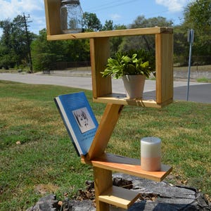 May include: A wooden, multi-tiered shelf unit with a natural finish. It holds a glass jar, a potted plant, a book, and a candle. The unit is outdoors, set on a tree stump, with a blue sky background.