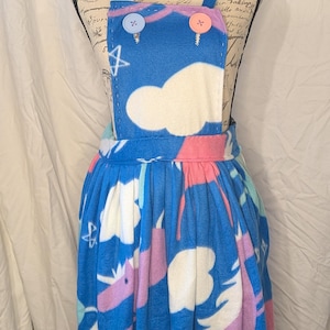 May include: Blue overall dress with a unicorn and cloud pattern. The dress features a blue top with two buttons and a pink trim. The skirt has a repeating pattern of unicorns in pink, purple, and blue.