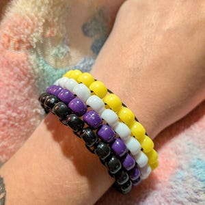 以下が含まれることがあります： 滑らかで丸いプラスチック製のポニービーズで作られた4連のブレスレットセット。ブレスレットは特定の色の順序で配置されており、黄色、白、紫、黒のビーズがそれぞれ1連ずつ並んでいます。