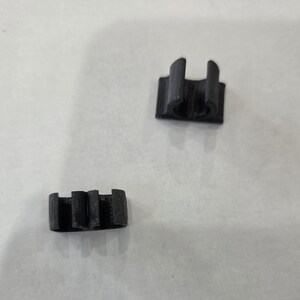 Peut inclure: Deux petits clips en plastique noir imprimés en 3D. L'un des clips a une ouverture circulaire et deux supports verticaux. L'autre clip a trois supports verticaux. Les deux clips sont sur une surface blanche.
