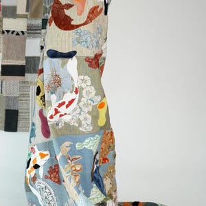 Könnte beinhalten: Eine große Patchwork-Decke mit einer Vielzahl von bunten Stoffen und Mustern, darunter Fische, Blumen und geometrische Muster. Die Decke ist über die Schultern einer Person gelegt.