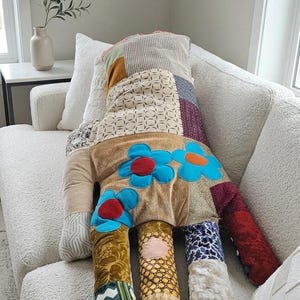 May include: A large, handmade decorative cushion in the shape of a figure, resting on a white sofa. The cushion features a patchwork design with various fabrics, including floral patterns and textured materials. Blue flower appliqués adorn the front.