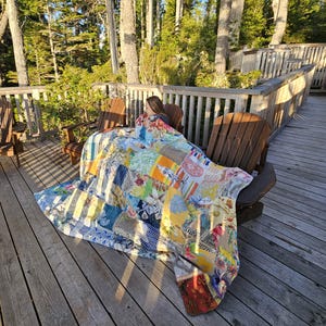 Op de afbeelding: Een kleurrijk patchwork-deken is over twee houten stoelen op een houten dek gelegd. Het deken is gemaakt van verschillende stoffen en patronen, waaronder strepen, bloemen en geometrische patronen. Het dek is omgeven door bomen en een houten railing.