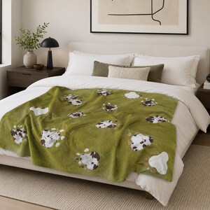 May include: A green throw blanket with a cow print design. The blanket features large, irregular shapes in white, brown, and black, scattered across the surface. The blanket is draped over a bed with white bedding, creating a cosy and inviting atmosphere.