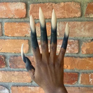 Puede incluir: Una mano con uñas largas y puntiagudas. Las uñas están pintadas con un efecto degradado, pasando de un beige claro en las puntas a un negro oscuro en la base. La mano está colocada contra una pared de ladrillos.