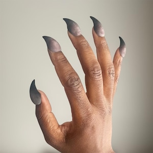 May include: A hand with long, dark gray, pointed fingernails. The nails are curved and extend beyond the fingertips. The skin tone is brown, and the background is a solid, light gray.