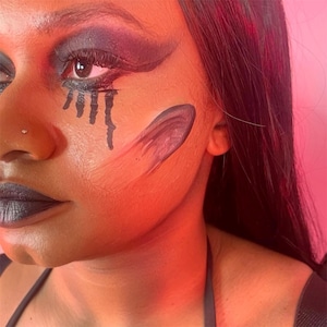 May include: Close-up of a face with dramatic makeup. Dark eyeshadow and lipstick are paired with black tear-like streaks under the eye and a curved design on the cheek. The skin has a warm, reddish tone.
