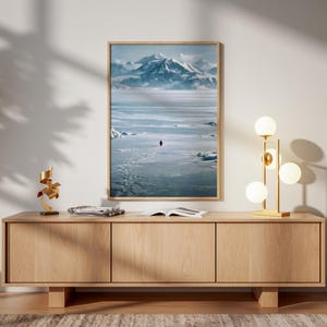 May include: A framed landscape print depicts a solitary figure traversing a snowy expanse towards snow-capped mountains. The print hangs above a light wood cabinet with a gold lamp and decorative sculpture.