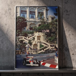 May include: Framed print of a Formula 1 race car speeding around a track in Monaco. The image features a red and white race car with the Marlboro logo, set against a backdrop of a grand building and lush greenery. The track has red and white striped barriers.