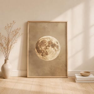 Op de afbeelding: Een rechthoekige wandkunstprint met een gedetailleerde, contrastrijke afbeelding van een volle maan gecentreerd tegen een effen, gedempte beige achtergrond. De print is ingelijst in een dunne, lichtgekleurde houten lijst en toont een realistisch maanoppervlak.