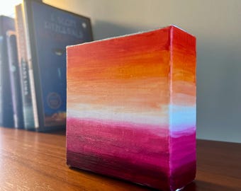 May include: A small, rectangular canvas art piece with a gradient of colors. The artwork transitions from deep magenta at the bottom to orange and red at the top, with a white and light blue band in the middle. The piece is displayed on a wooden surface.