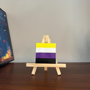 Może przedstawiać: Mały obraz na płótnie na drewnianym sztaludze, przedstawiający flagę dumy osób niebinarnych. Flaga ma poziome paski w kolorze żółtym, białym, fioletowym i czarnym. Sztaluga jest wykonana z jasnego drewna, a obraz jest eksponowany na ciemnej drewnianej powierzchni.