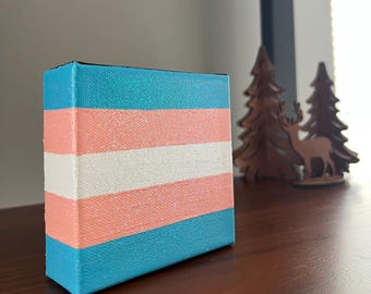 May include: A small canvas with a transgender pride flag painted on it. The flag is made of light blue, pink, and white stripes with a glittery finish.