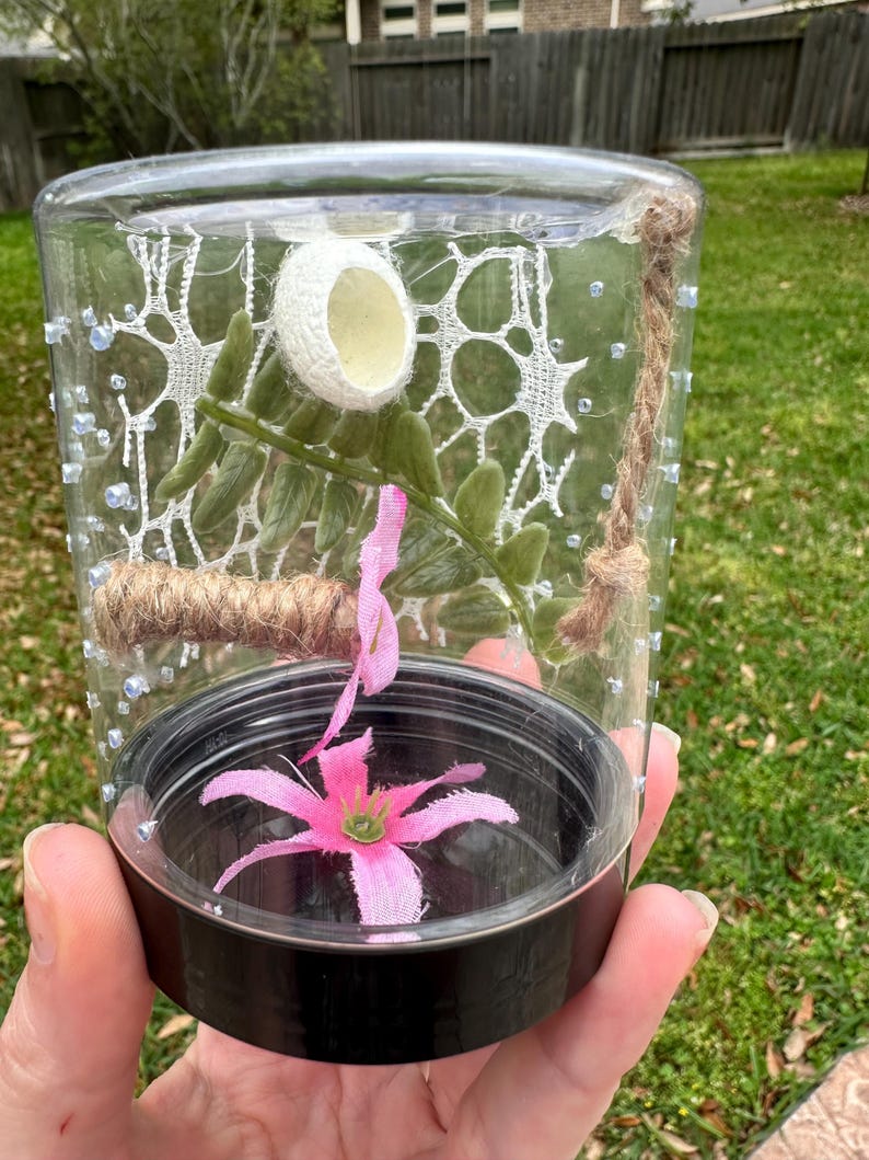 Puede incluir: Un tarro de vidrio transparente con tapa negra contiene una servilleta de encaje blanca, un peque&ntilde;o huevo blanco, una rama verde con hojas, una cuerda marr&oacute;n y una flor de tela rosa.