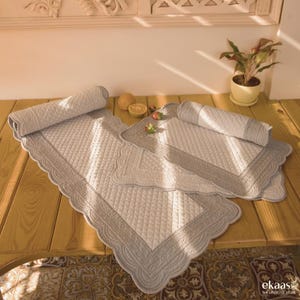 May include: Two quilted table runners in a light grey and white colour scheme with a scalloped edge. The runners are rolled up and placed on a wooden table.