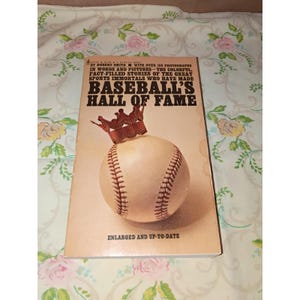 May include: A vintage book titled "Baseball's Hall of Fame" with a baseball and crown illustration. The cover is beige with black and brown text. The book includes over 150 photographs and stories of baseball greats. The text reads "Enlarged and Up-to-Date."