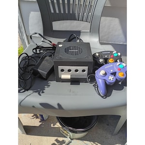 May include: A black Nintendo GameCube video game console with a power adapter, a black controller, and a purple controller. The console features a front panel with four controller ports and a memory card slot, with the GameCube logo on the top lid.