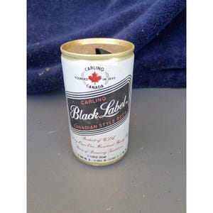 May include: A vintage Carling Black Label beer can. The can is white with black and gold accents, featuring the Carling logo and the words "Black Label" in a stylized font. The can is 12 fluid ounces (355ml).