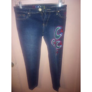 May include: Dark wash denim jeans with a colorful embroidered design on the left leg. The jeans have a classic five-pocket design and a button and zipper closure. The word "COOL" is embroidered on the waistband.