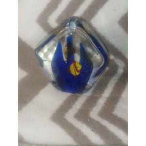 May include: A clear glass paperweight with a blue and yellow fish design. The paperweight has a square shape with rounded edges. The fish are yellow with red accents and are set against a blue background.