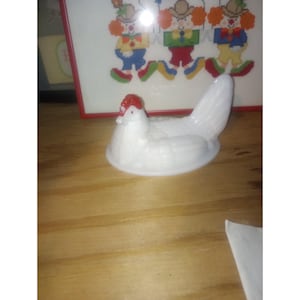 May include: A white ceramic hen-shaped dish with a red comb. The dish is on a wooden surface, with a framed cross-stitch picture of clowns in the background. The hen is a decorative item.