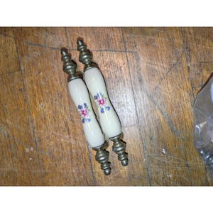 May include: Two elongated cabinet pulls featuring a smooth, cream-colored ceramic center section decorated with a small floral pattern of pink and blue flowers. The ends of each handle are finished with a metallic, antique-style brass hardware.