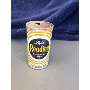 Peut inclure: Une canette de bière vintage Light Reading Premium. La canette cylindrique présente des rayures blanches et dorées. L'étiquette affiche "Light Reading" dans une police stylisée, avec "Premium Beer" en dessous. La canette est légèrement rouillée.
