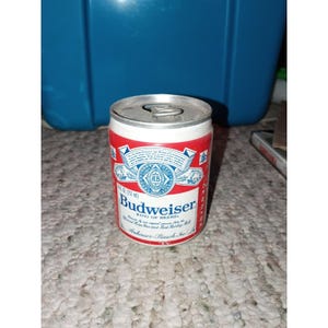 Peut inclure: Une canette de bière Budweiser vintage, rouge et blanche avec le logo et le texte Budweiser. La canette est cylindrique avec un dessus argenté à tirette. La canette mesure environ 9 cm de haut.
