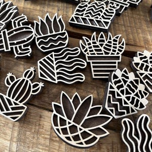 May include: A set of ten wooden plant cutouts in black and white. The cutouts are in the shape of various plants, including cacti, succulents, and ferns. The cutouts are all different sizes and shapes, and they are all made of a dark wood.