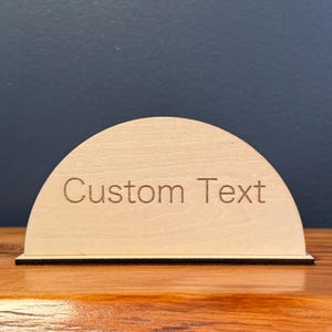 May include: A light-coloured wooden sign in a semi-circular shape, with the words "Custom Text" engraved on the front. It has a black base and is placed on a wooden surface. The background is a dark blue wall.