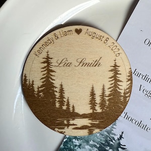 May include: A circular wooden wedding favour with engraved text and a forest scene. The text reads "Kennedy & Liam", a heart symbol, "August 8, 2026", and "Lia Smith". The design features trees and a lake.