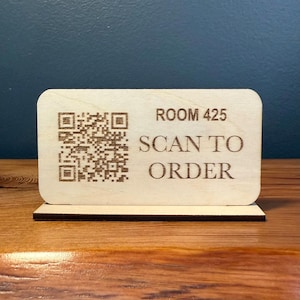 Peut inclure: Panneau de table en bois clair avec coins arrondis et base rectangulaire. Il présente un code QR et le texte "ROOM 425 SCAN TO ORDER" gravé.