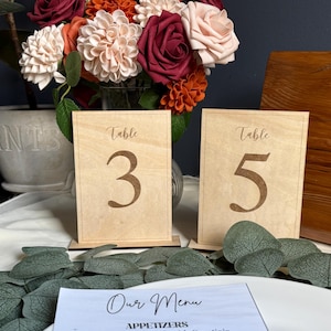 May include: Two wooden table number signs, labelled "Table 3" and "Table 5", with a floral arrangement in the background. The flowers are in shades of red, orange, and cream. A menu with the text "Our Menu" is visible.