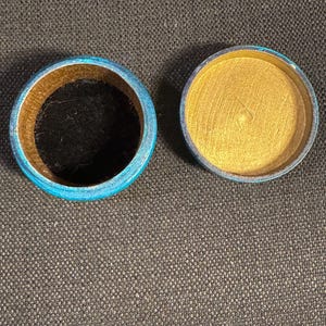 May include: Open round wooden box with a blue rim and gold interior. The box is open, revealing a black interior in the base. The lid is separate, showing a gold interior. Likely for jewellery or small items.