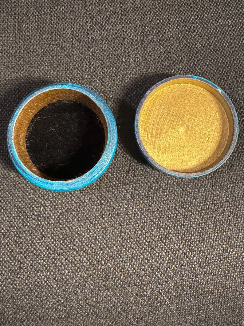 May include: Open round wooden box with a blue rim and gold interior. The box is open, revealing a black interior in the base. The lid is separate, showing a gold interior. Likely for jewellery or small items.