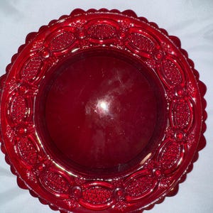 May include: A vibrant red glass plate with a decorative, raised pattern around the rim. The plate has a scalloped edge and a smooth, reflective center. This vintage-style plate is suitable for serving or display.