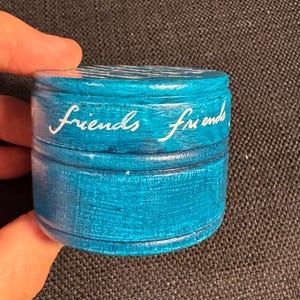 May include: A small, round, turquoise-painted wooden box with the word "friends" in white script on the side. The box has a lid and appears to be handmade. The box is held in a person's hand.