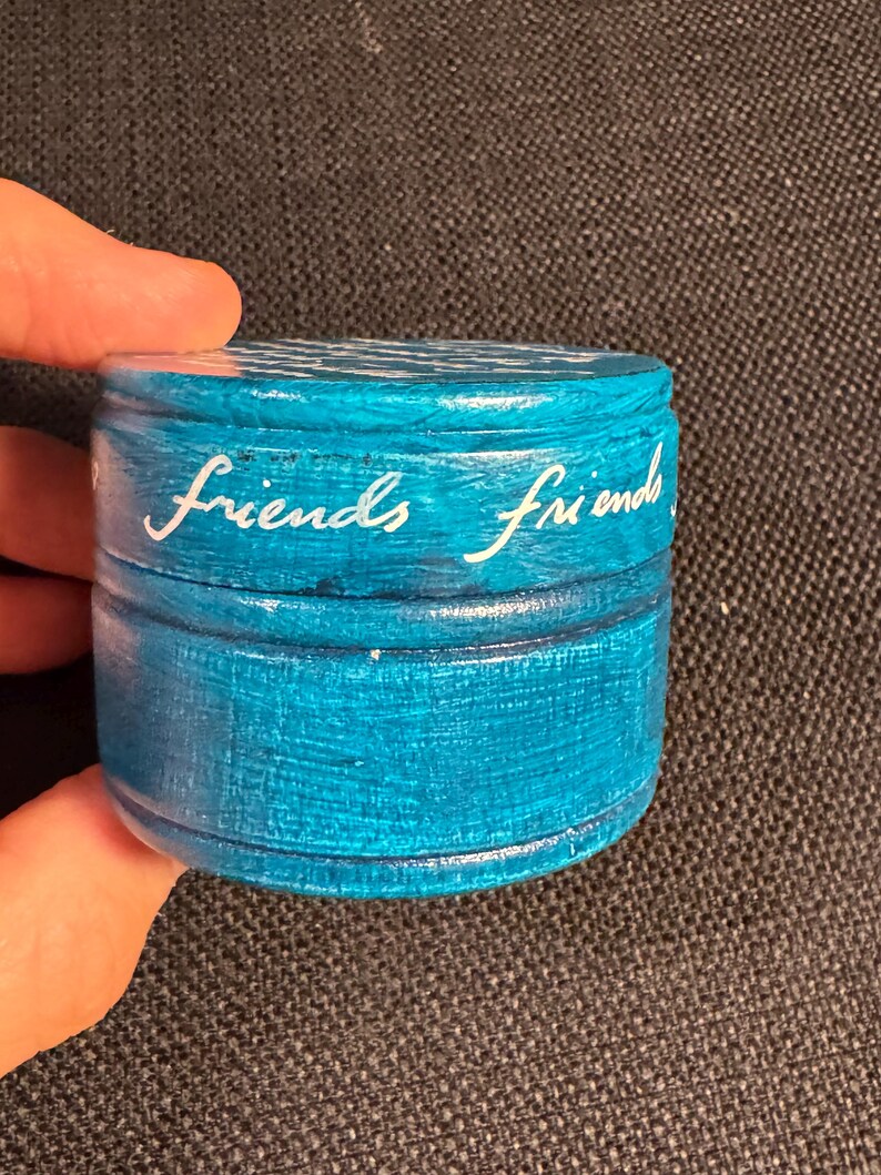 May include: A small, round, turquoise-painted wooden box with the word "friends" in white script on the side. The box has a lid and appears to be handmade. The box is held in a person's hand.