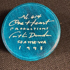 May include: A circular, turquoise object with white lettering. The text reads "No. 614," "One Heart Productions," "Caitlin Dunda," "Seattle WA," and the year "1997." The object is displayed on a dark, textured surface.