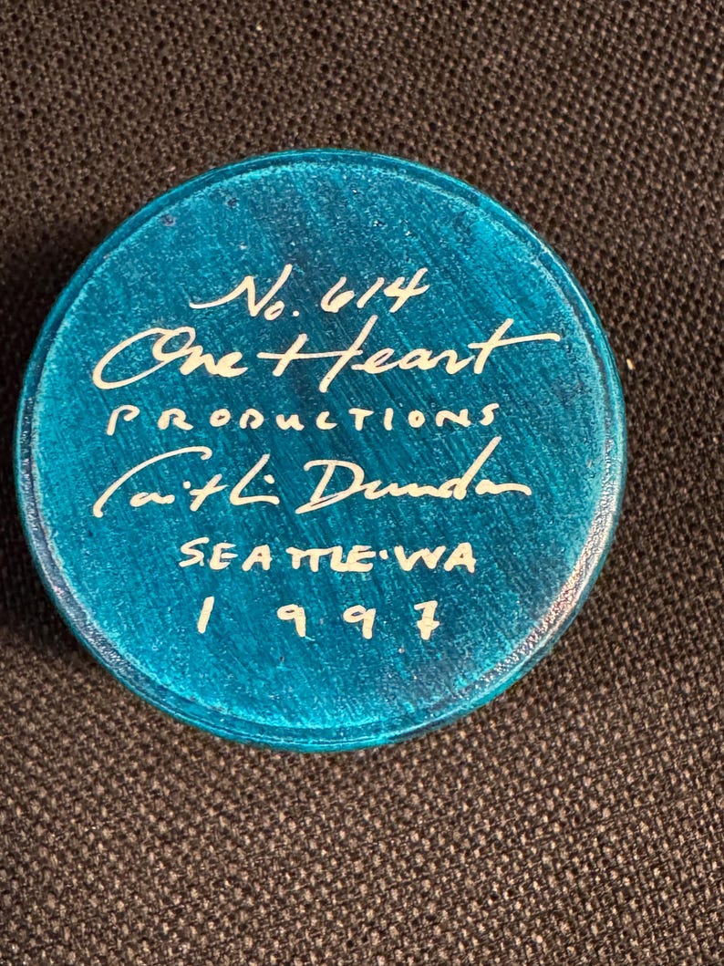 May include: A circular, turquoise object with white lettering. The text reads "No. 614," "One Heart Productions," "Caitlin Dunda," "Seattle WA," and the year "1997." The object is displayed on a dark, textured surface.