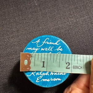 May include: A round, turquoise button with white script reading "A friend may well be..." and "Ralph Waldo Emerson." The button is approximately 5 cm in diameter. A measuring tape is placed across the button.