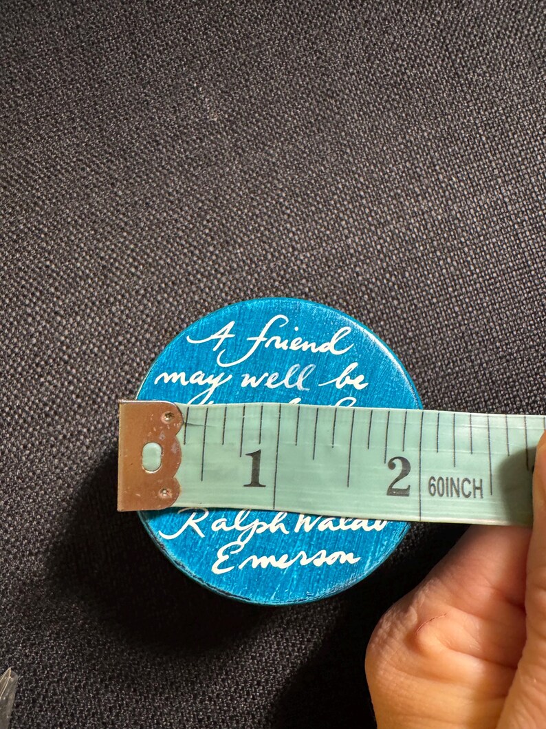 May include: A round, turquoise button with white script reading "A friend may well be..." and "Ralph Waldo Emerson." The button is approximately 5 cm in diameter. A measuring tape is placed across the button.