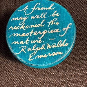 May include: A circular, teal wooden item with white cursive writing. The text says, "A friend may well be reckoned the masterpiece of nature. - Ralph Waldo Emerson."