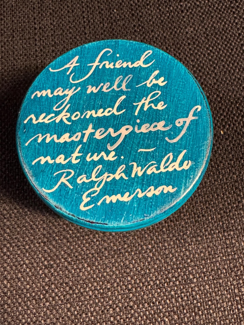 May include: A circular, teal wooden item with white cursive writing. The text says, "A friend may well be reckoned the masterpiece of nature. - Ralph Waldo Emerson."
