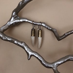 May include: A pair of earrings made from antler and bullet casings. The antler is white and the bullet casings are gold. The earrings are hanging from a silver branch.