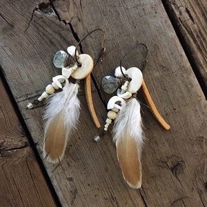May include: A pair of dangle earrings with white feathers, white beads, and light blue stones. The earrings are made with a brown wire and have a hoop design.
