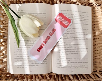 以下が含まれることがあります： 赤いチケットのようなデザインが上部にある、ピンク色の「SMUT BOOK CLUB」の文字が入ったブックマークが、白いチューリップと開いた本に挟まれています。