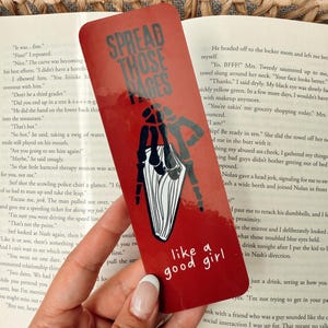 May include: Red bookmark with a black illustration of a hand holding a book. The bookmark has the text "Spread Those Pages Like a Good Girl" printed on it.