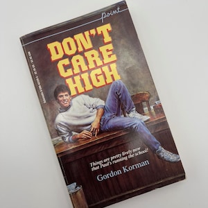 May include: A paperback book titled "DON'T CARE HIGH" with a cover illustration of a young person reclining on a desk. The book has yellow and orange lettering and the author's name is Gordon Korman. The book is a "Point" book.